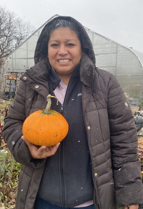 A woman stands in a garden wearing a winter coat and holding an orange pumpkin. 